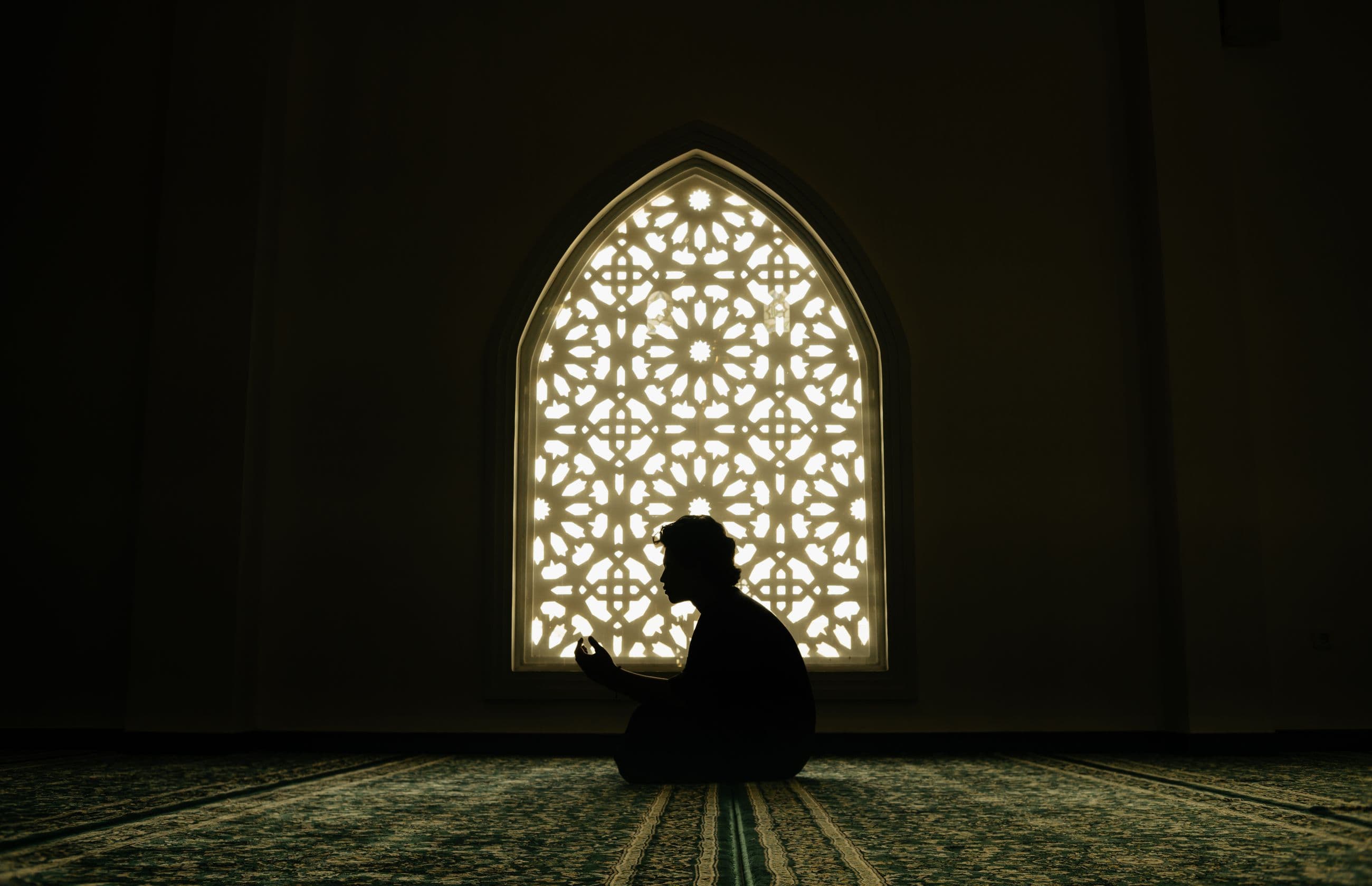 A person sitting in silhouette with hands raised in dua, in front of a lit arched lattice window in a mosque.